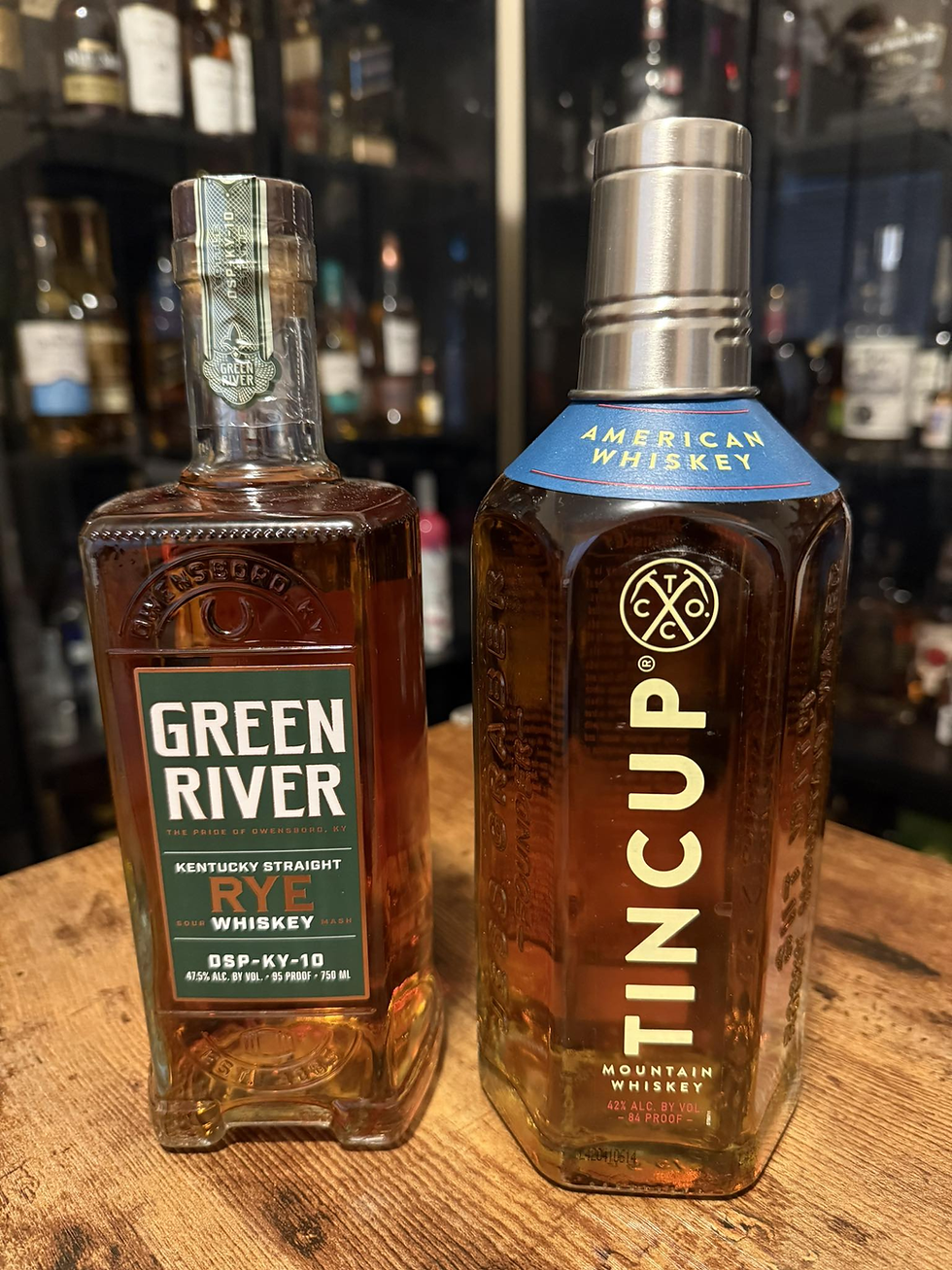 Two whiskey bottles on a wooden table, Green River Rye and Tincup Mountain Whiskey, with blurred bar shelves behind.