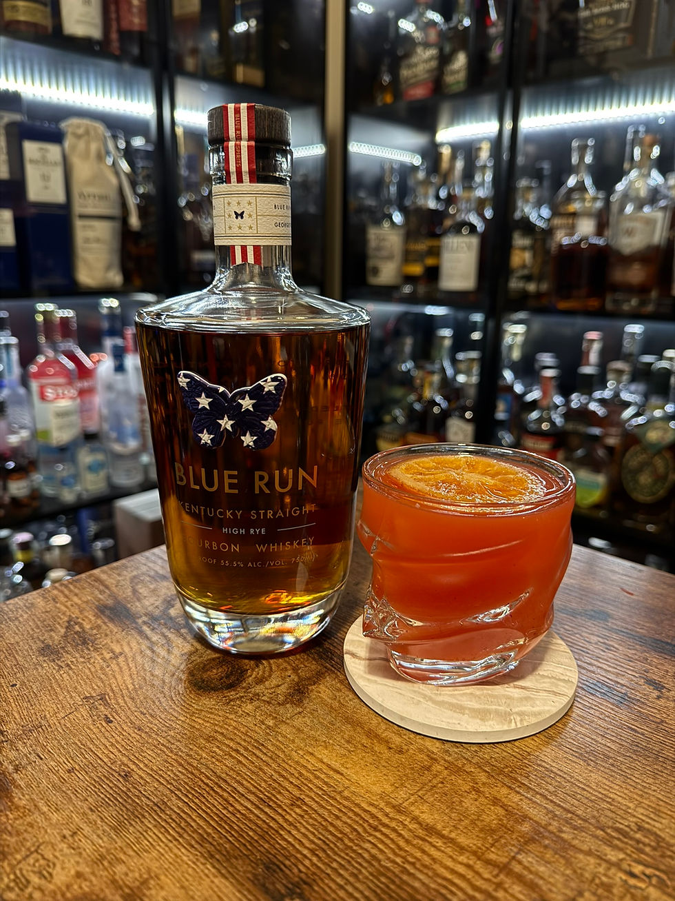 Blue Run bourbon bottle beside an orange cocktail on a wooden bar, with blurred liquor shelves in the background.