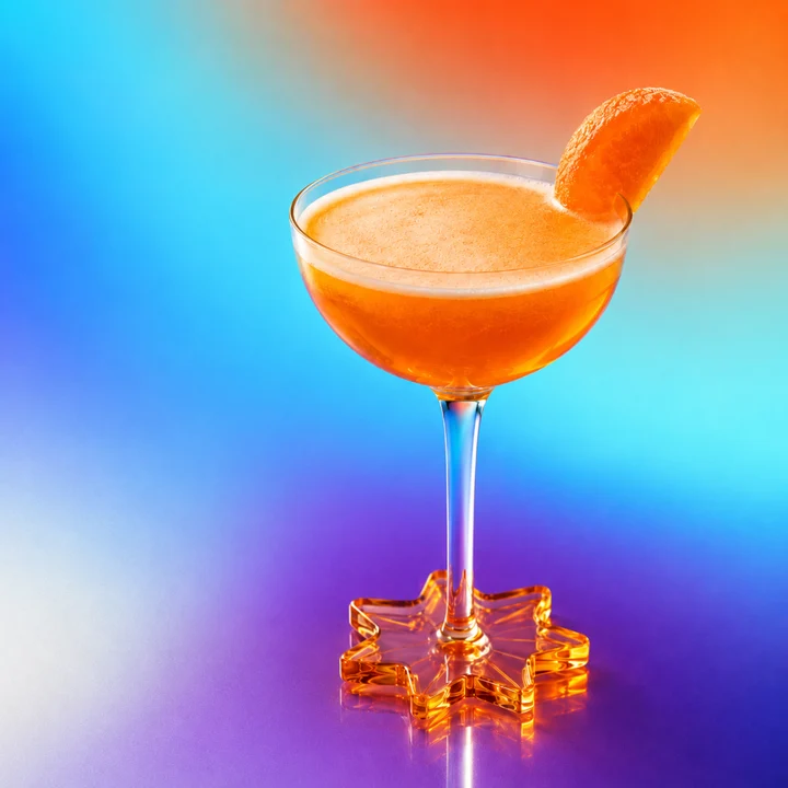 Orange cocktail in a coupe glass with an orange slice on a star-shaped amber base, against a bright blue-orange gradient background.