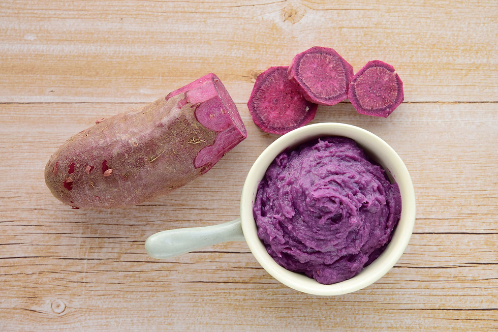 Purple sweet potato, sliced rounds, and a bowl of purple mash on a light wooden tabletop.
