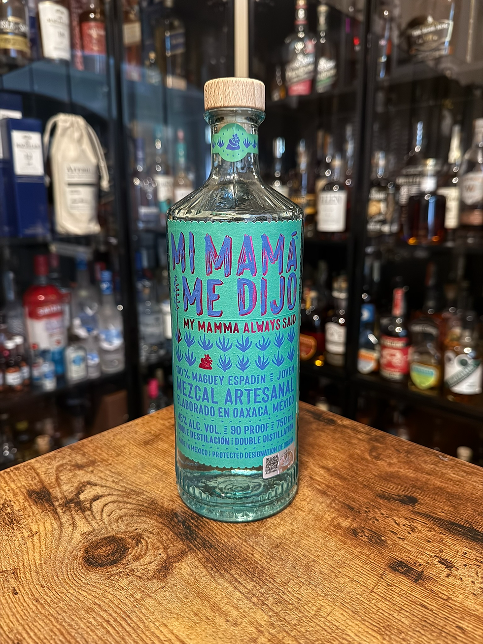 Turquoise Mi Mama Me Dijo mezcal bottle on a wooden table, with blurred liquor shelves behind it in a bar-like setting.