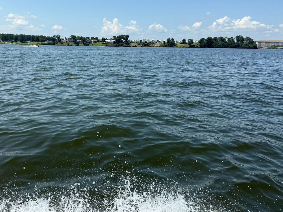 Boat wake splashing on a lake, with a distant tree-lined shore, houses, and a blue sky with fluffy clouds.