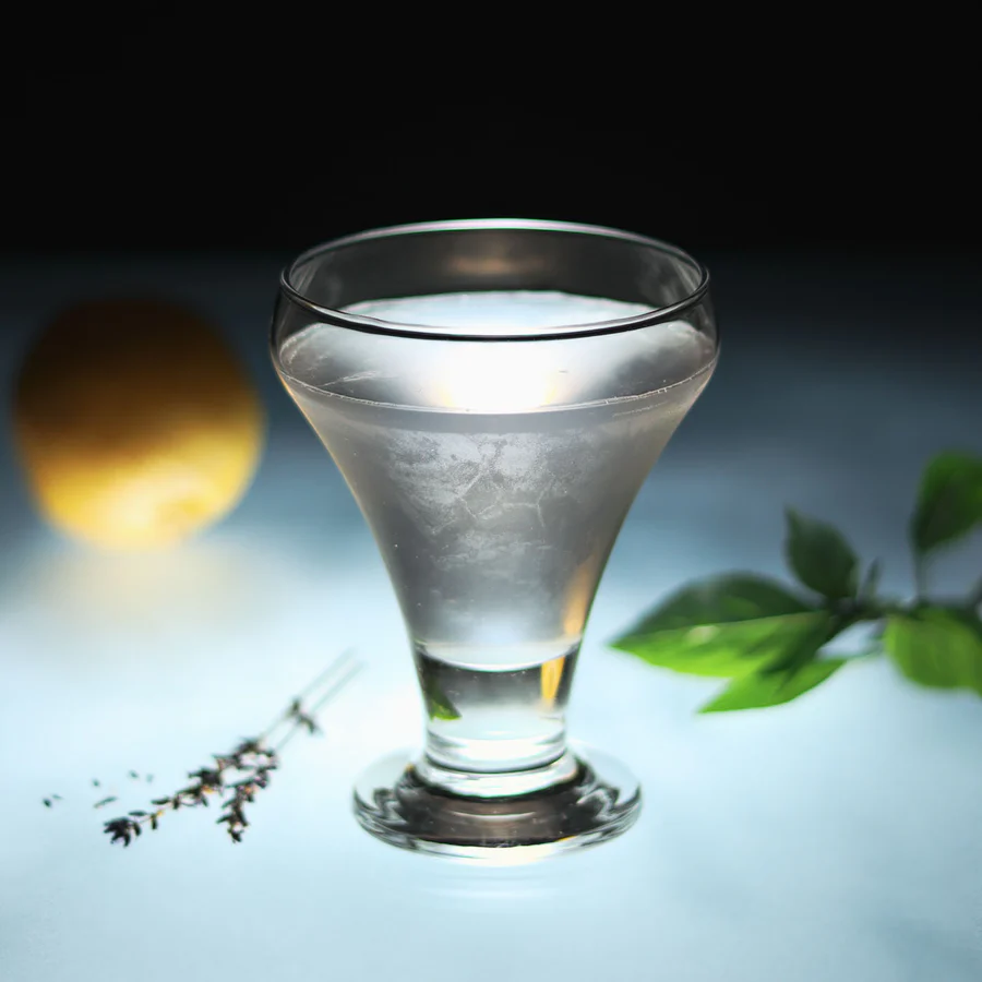 Clear cocktail glass filled with chilled water on a blue surface, with herb sprig and green leaves against a dark background