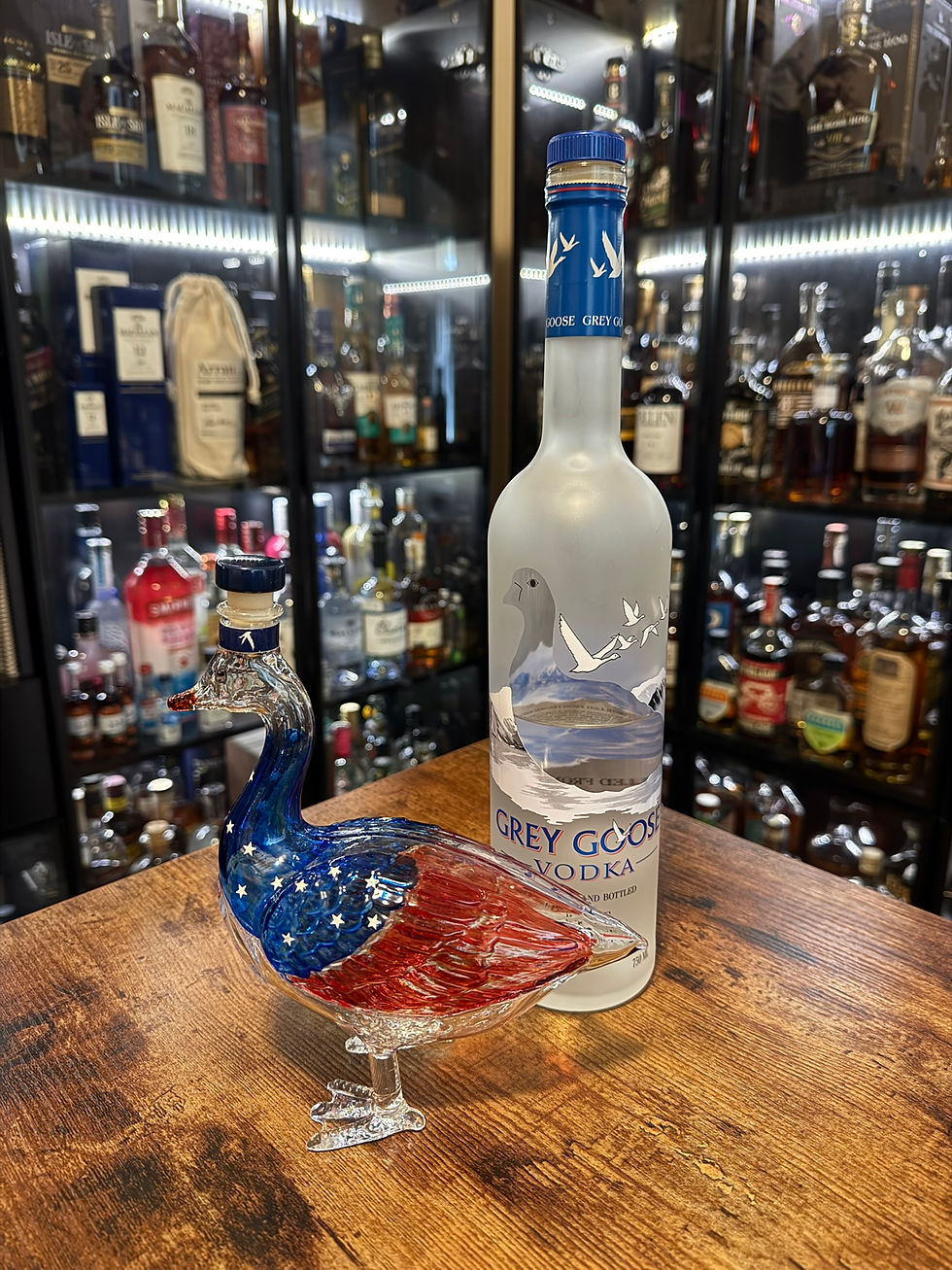 Grey Goose vodka bottle beside a red, white and blue glass goose on a wooden table, with liquor bottles lit in the background.