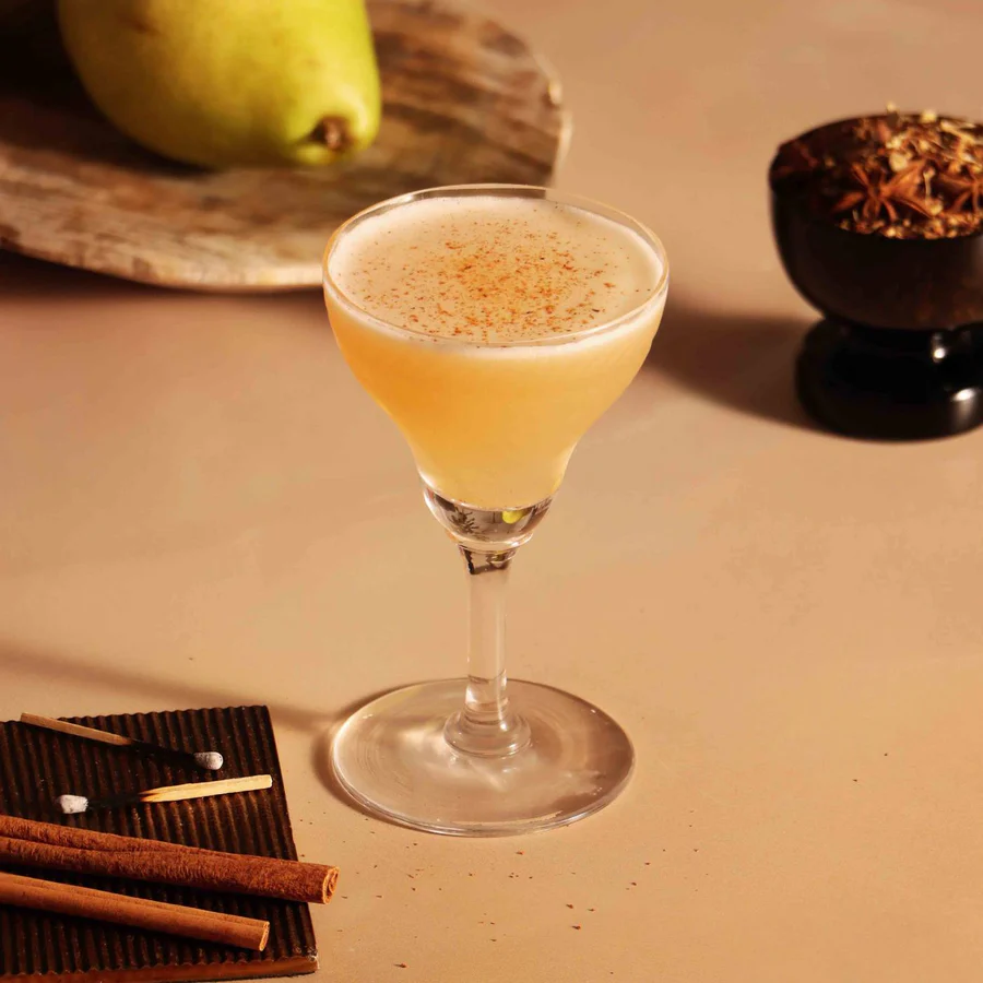 Pear cocktail in a coupe glass dusted with spice, beside a pear, cinnamon sticks, and star anise on a warm beige table.
