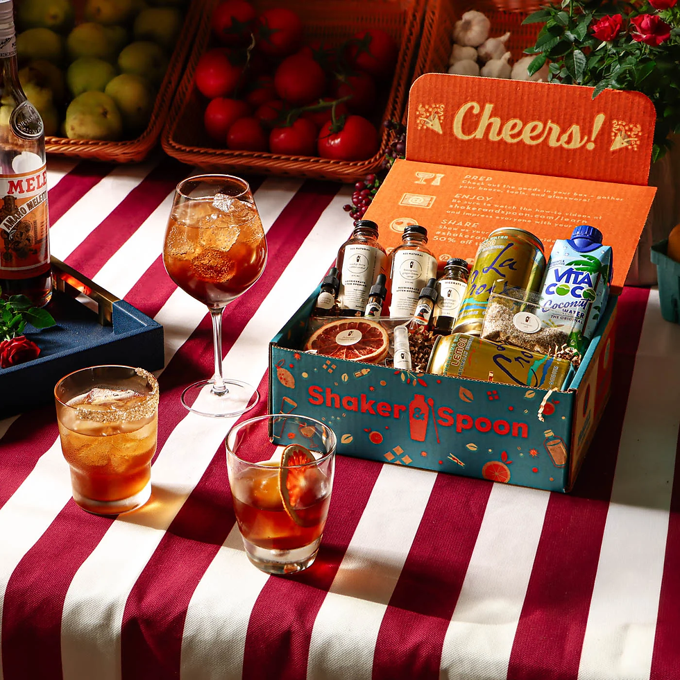 Cocktail gift boxes labeled Shaker & Spoon and Cheers! sit on a red-and-white striped table with drinks and fruit baskets.