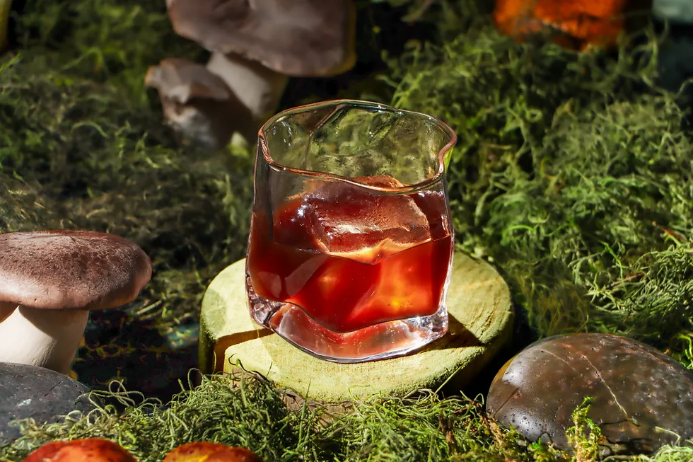 Glass of red iced drink on a mossy forest floor, surrounded by mushrooms, in warm sunlight