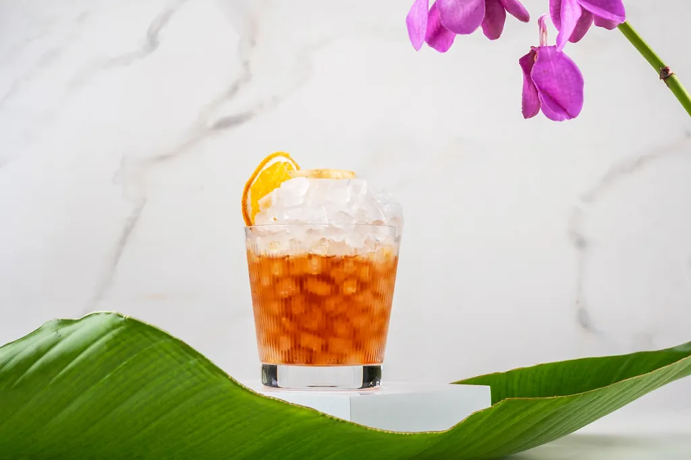 Iced orange cocktail in a glass with orange slice, framed by green leaves and purple orchid petals on a white marble background.