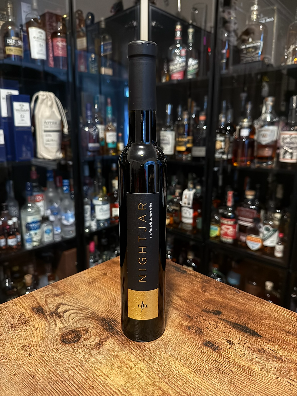 Black NIGHTJAR dessert wine bottle on a wood table, centered before a blurred wall of liquor bottles in a dim cellar.