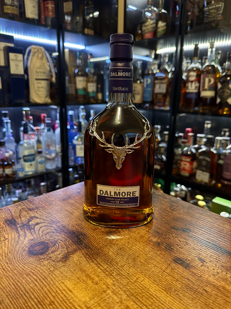 The Dalmore 12-year Scotch bottle on a wooden table, with a backlit whiskey shelf and blurred liquor bottles behind it.