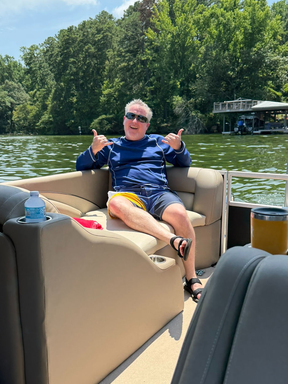 Smiling man in sunglasses on a pontoon boat gives shaka signs on a sunny lake with trees and docked boats.