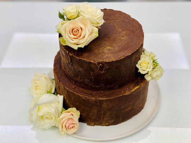Tort cu două etaje, acoperit în ganache de ciocolată neagră și decorat cu flori naturale crem și roz pal, realizat la Noeme București pentru o aniversare în familie.