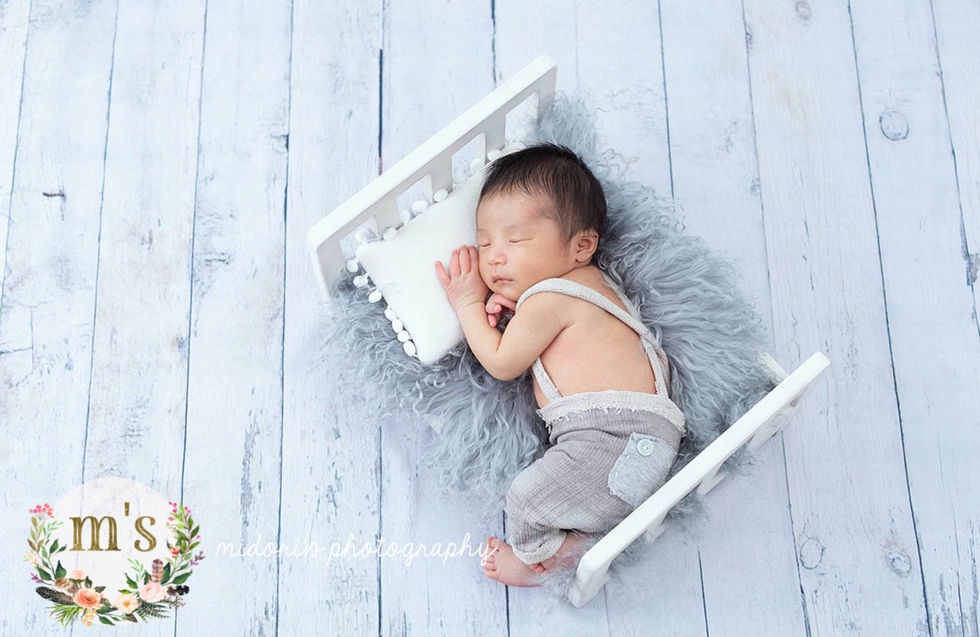 白いミニチュアベッドでうつ伏せに眠る新生児。ベージュのサロペットとグレーのファーを合わせたニューボーンフォト