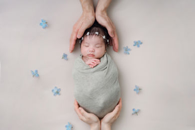 ご両親の手にそっと包まれ、グレーのおくるみで眠る新生児。まわりに青い小花