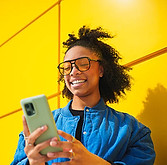 A smiling young woman with curly hair wearing glasses and a blue jacket, looking at her smartphone against a vibrant yellow background.
