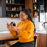 A woman with braided hair wearing a yellow sweater sitting at a kitchen counter, holding a smartphone and a payment card, seemingly engaged in an online transaction.