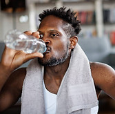 A man wearing a white tank top and a towel around his neck drinking from a clear water bottle after a workout.