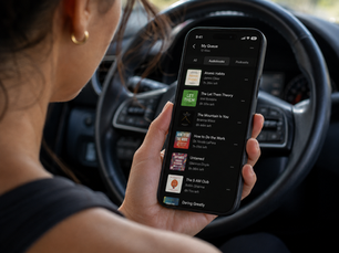 A woman in workout clothes sits in her car scrolling through a queue of self-help and personal growth audiobooks on her phone before heading to the gym.