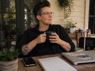 A non-binary adult sits comfortably on a back patio holding a coffee mug, leaving their phone, laptop, and calendar untouched on the table.