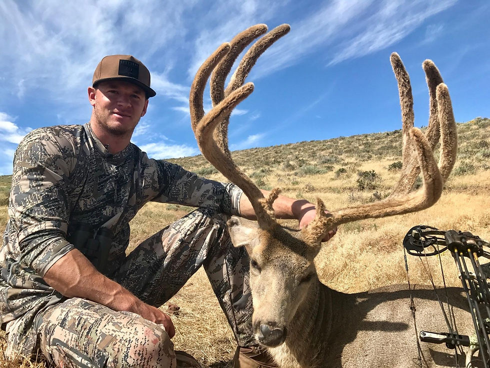 A man in camouflage hunting gear crouching beside a deer with large antlers in a dry, grassy field.