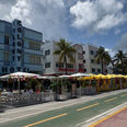 Colorful beachfront hotels and restaurants with palm trees and outdoor café seating under a bright cloudy sky along a quiet street