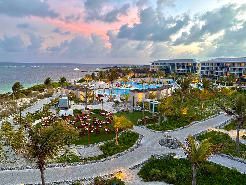 Tropical beachfront resort at sunset with palm trees, pool, and ocean under a pink-blue cloudy sky.