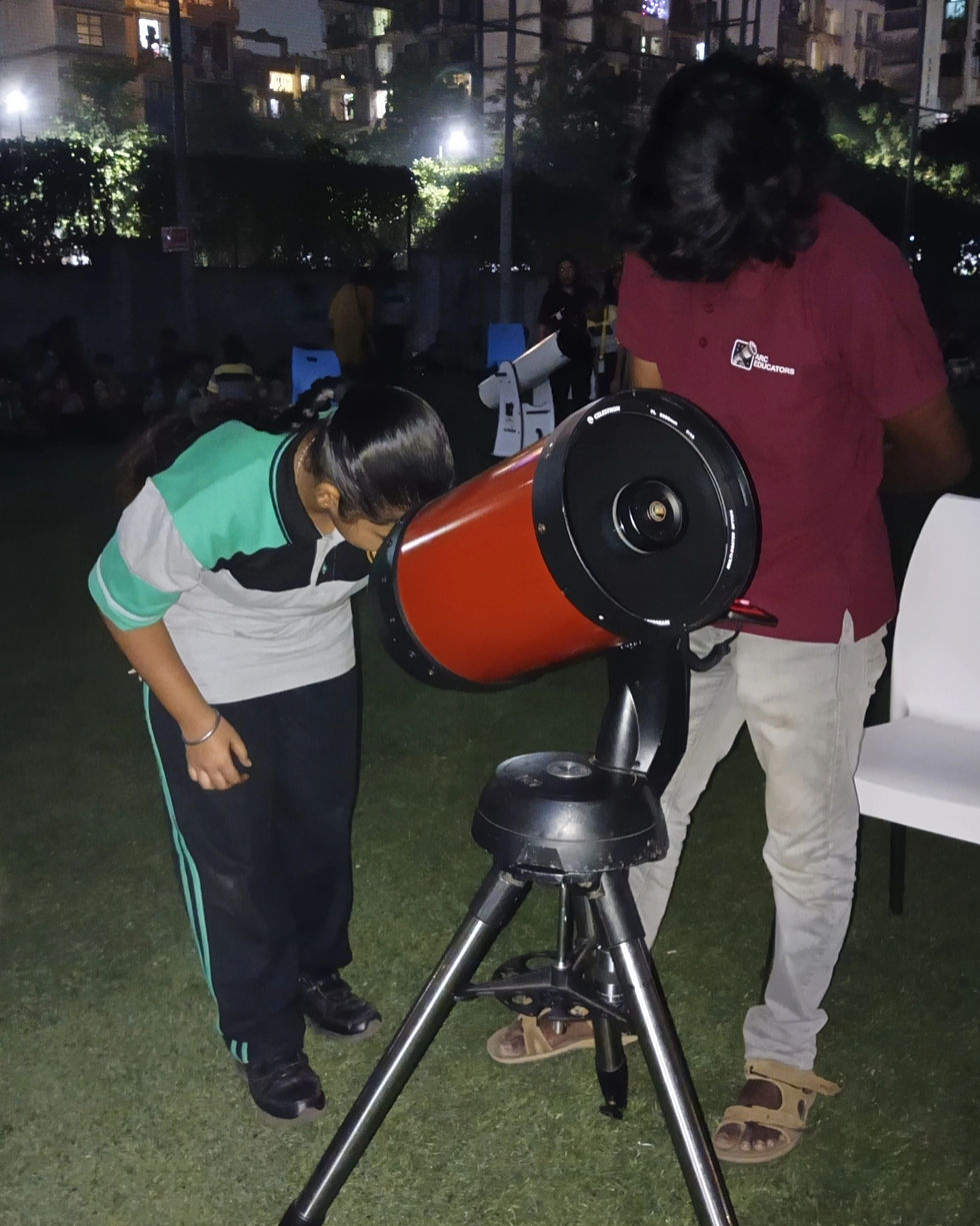 A person in a green and white shirt looks into a telescope at night on grass. Another person stands nearby. Buildings lit in the background.