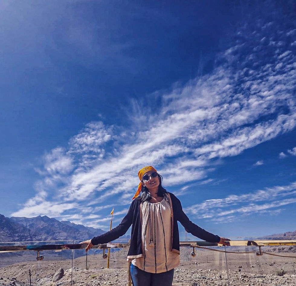 Woman in sunglasses and orange bandana smiles, arms outstretched, against a clear blue sky with wispy clouds and distant mountains.