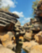 Eroded sandstone canyon with rugged orange-brown rocks, sparse vegetation, and bright blue sky with fluffy clouds, creating a serene mood.