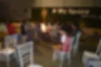 People sit around a campfire at night in a grassy area. Chairs encircle them. Background shows "# My Space HOTELS" on a wall.