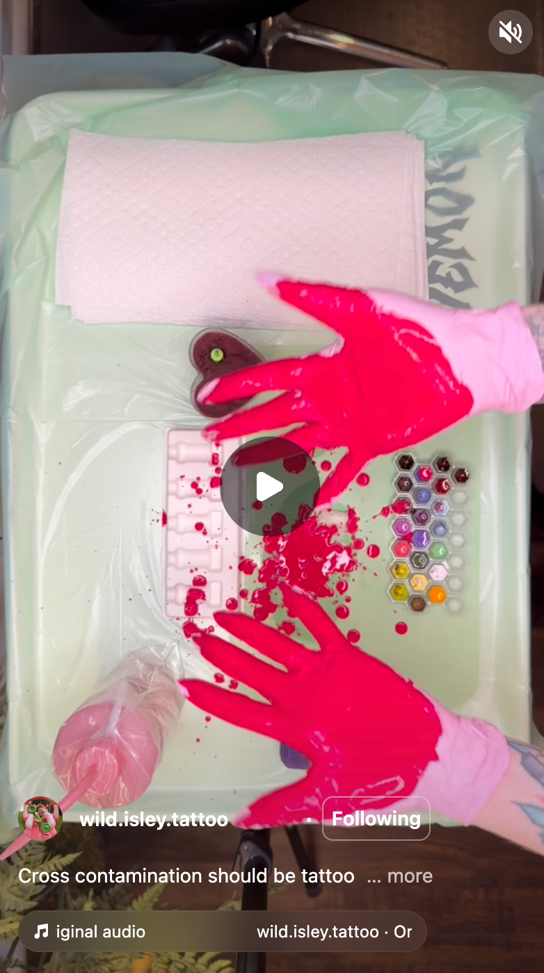 Overhead view of a tattoo artist wearing bright pink gloves, demonstrating sanitation practices with red liquid on a covered workstation. The setup includes paper towels, ink caps, and disinfected tools. The video shows how cross-contamination can occur and emphasizes proper hygiene in tattooing.
