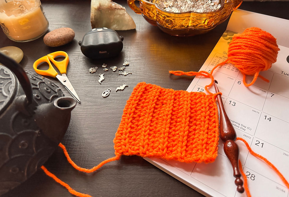 Orange crochet square with wooden crochet hook and calendar in background, next to a cast icon tea kettle and yellow scissors