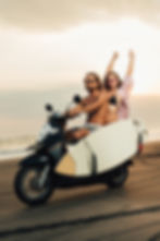 boyfriend riding scooter with surfboard on beach, girlfriend sitting with raised hands.jpg