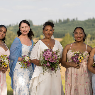 Five smiling women, bride and bridesmaids, holding bouquets outdoors.