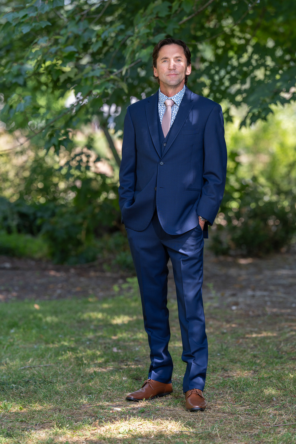 Man in blue suit standing confidently outdoors among green foliage.