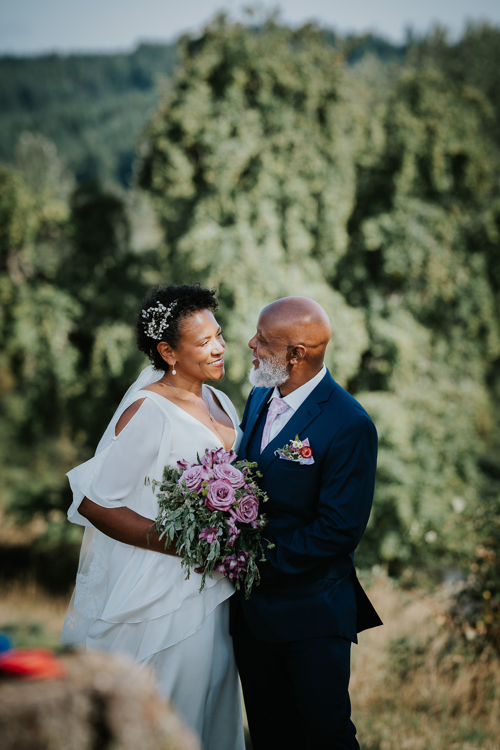 Happy wedding couple gazing lovingly, holding purple bouquet, intimate farm wedding.