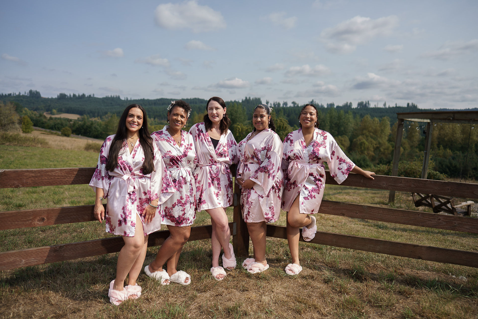 Five bridesmaids in floral robes and slippers pose outdoors on a farm.