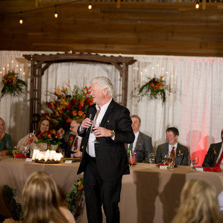 Man in suit gives speech at indoor wedding reception, guests listening.