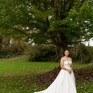 Smiling bride in white strapless gown stands under a large tree.