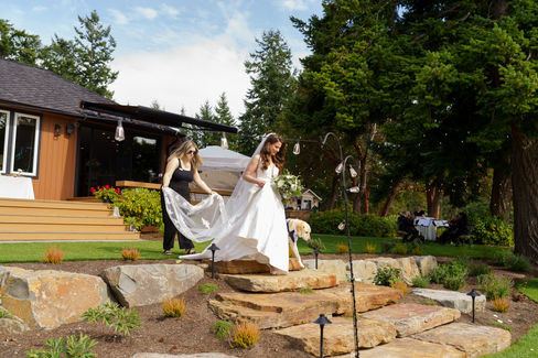 Wedding assistant helps bride descend outdoor stone steps in white gown.