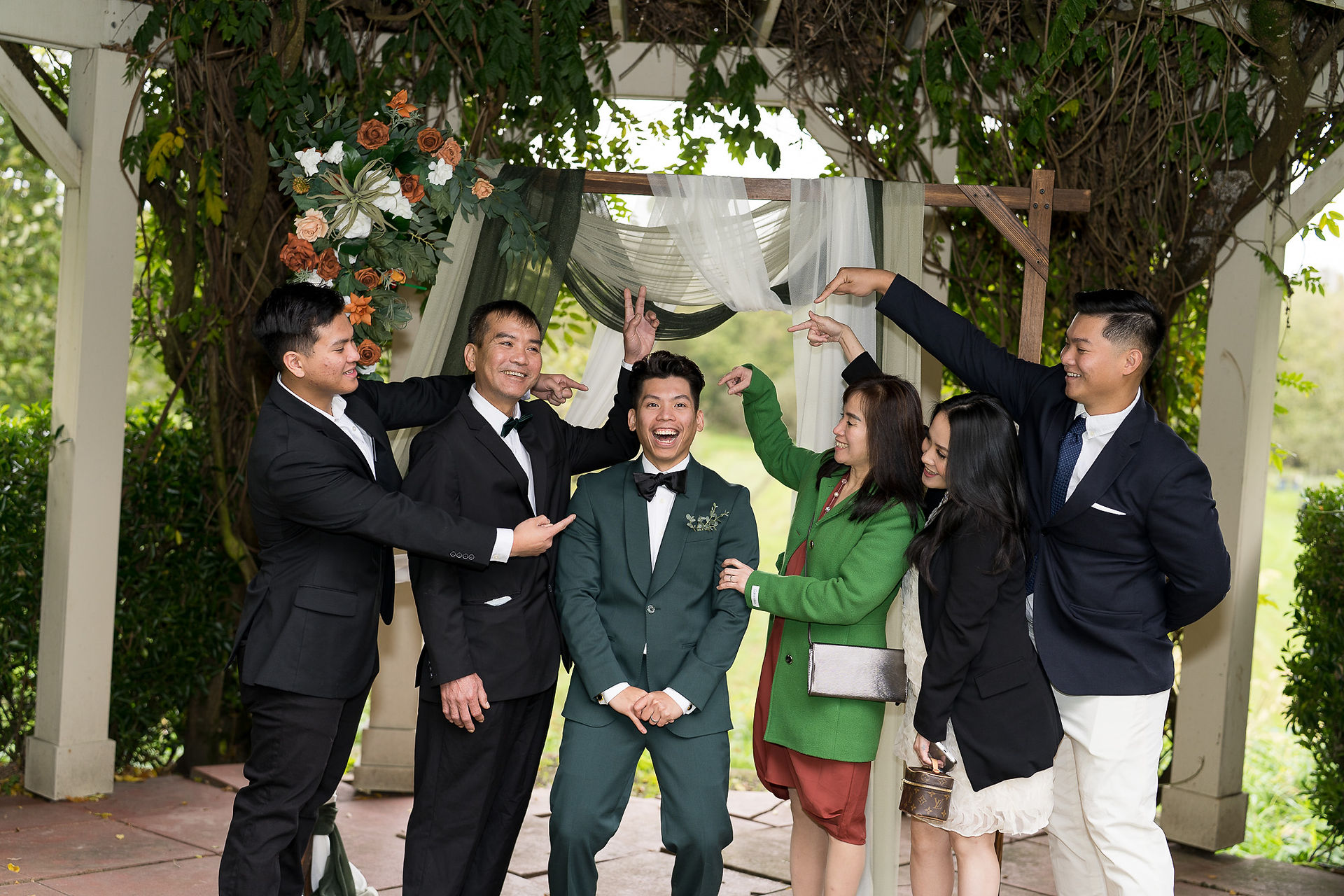 Happy groom in green suit with five smiling guests pointing.