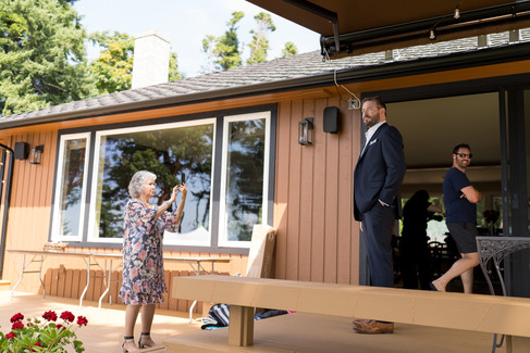 Woman photographs man in suit on backyard deck beside house. Formal event.