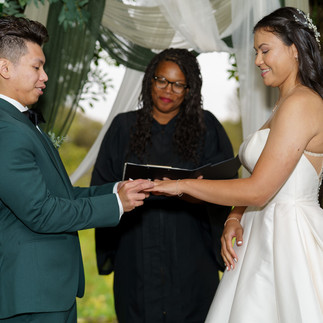 Groom placing ring on bride's finger during outdoor wedding ceremony with officiant.