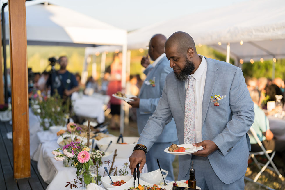 Groomsman in light blue suit serving food at outdoor wedding reception.