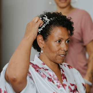 Woman adjusting white floral hairpiece, wearing a floral robe and pearl earrings.