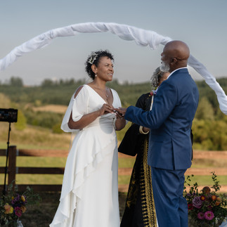 Bride placing ring on groom's finger under white arch outdoors.
