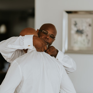 Father helping groom adjust white shirt collar before wedding ceremony.