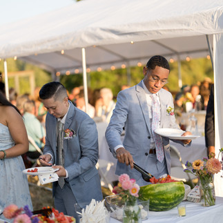 Two well-dressed men serving fruit from an outdoor wedding buffet.