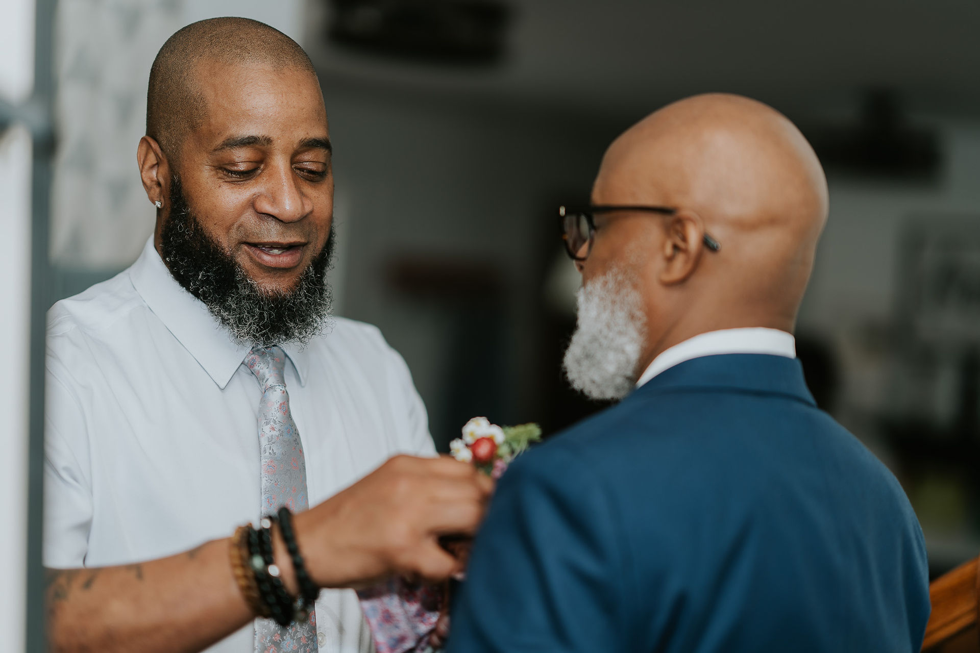 Man with beard pinning boutonnière on another man's suit jacket.