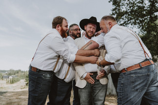 Groomsmen joyfully lift groom in black hat outdoors, celebrating wedding.